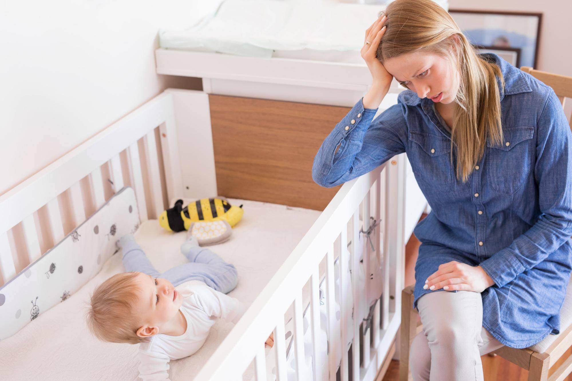 tired mom resting elbow on crib with baby in it